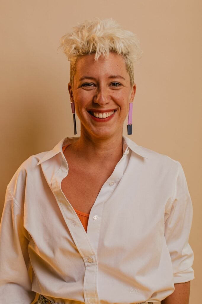 Photo d'une femme souriante avec une coiffure courte et blonde, portant une chemise blanche.